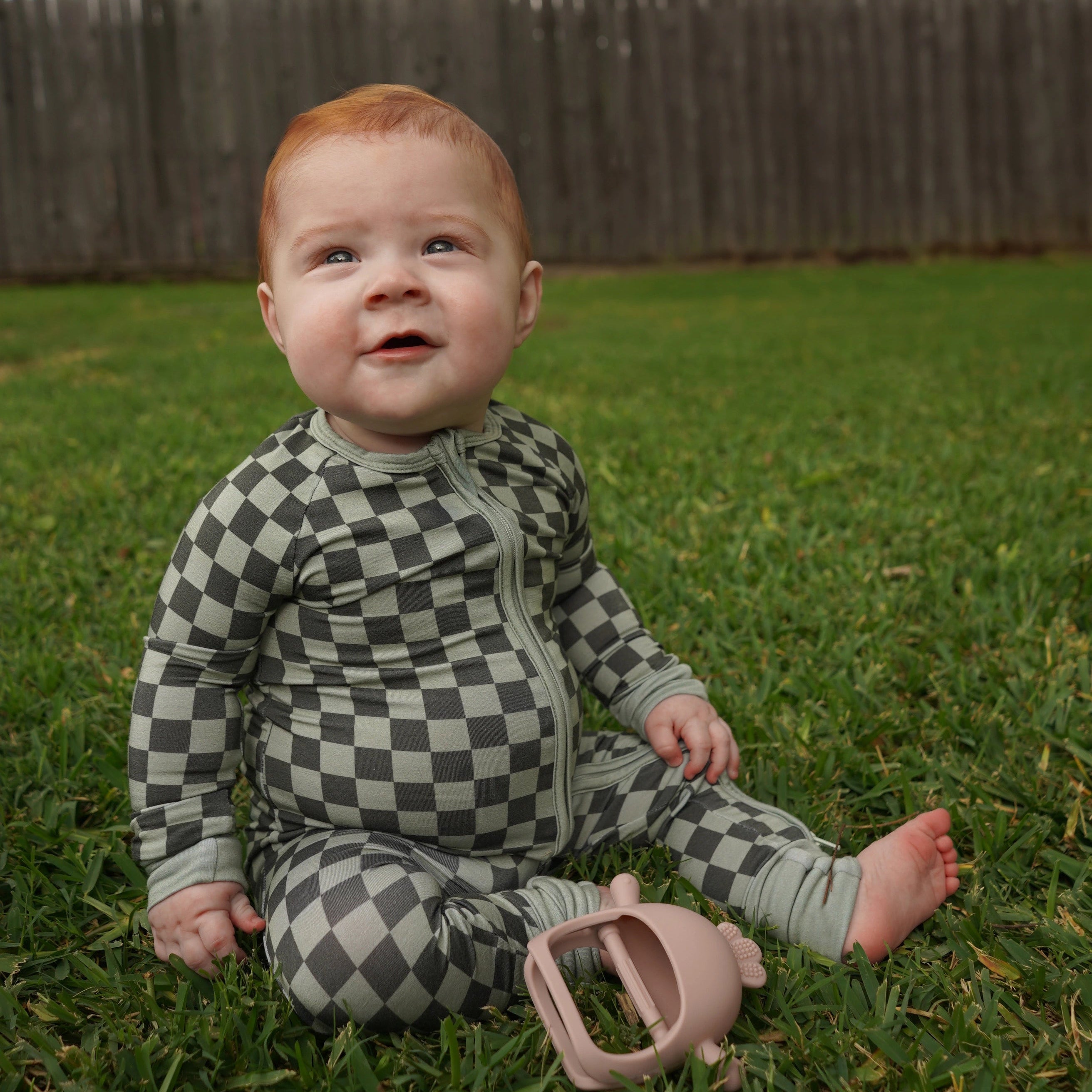 Baby in evergreen zippy sitting in field of grass outside.