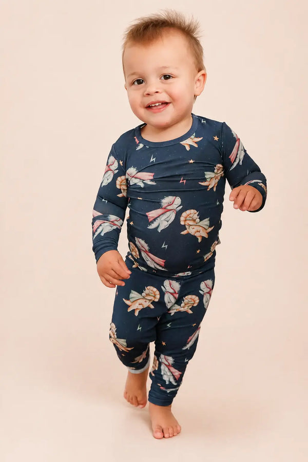 A smiling toddler walking barefoot against a solid light beige background, wearing a navy blue long-sleeve pajama set with a whimsical print of fairies, stars, and butterflies.